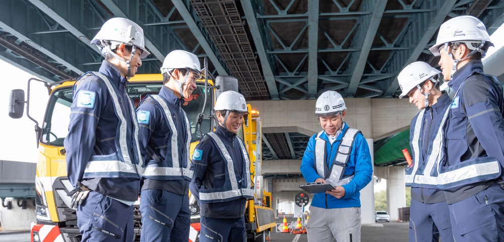 高速道路の高架下で責任者を囲み打ち合わせをするスタッフたち