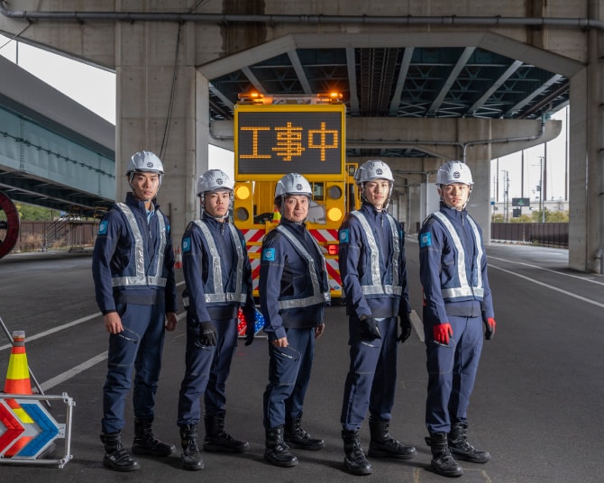 「工事中」表示の車の前で整列する5名の警備スタッフ