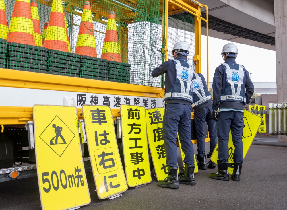 高速道路の高架下で工事標識やカラーコーンを積んだ作業車の前に立つ警備スタッフ